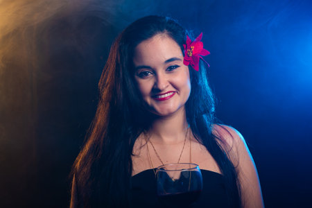 Beautiful woman with long black hair with a red flower on her left ear smiling and looking at the camera. Studio photo with a smoky colored background.の写真素材