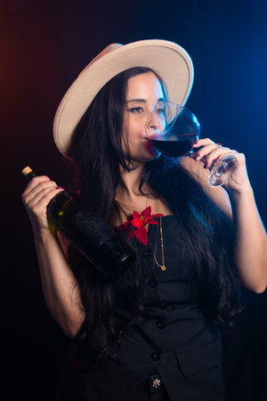 Beautiful woman wearing brown hat holding a bottle and drinking wine from the glass. Studio photo with smoky colorful background.の写真素材