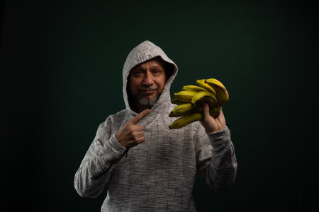 Mysterious man wearing hoodie holding bunch of yellow bananas. Isolated on dark background.の写真素材