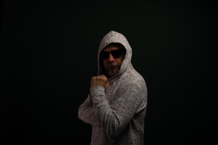 Portrait of man wearing sunglasses and hooded sweatshirt standing, motionless, making hand gestures. Isolated on dark background.の写真素材