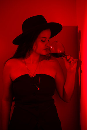 Portrait of beautiful woman in black clothes and hat drinking wine from a glass leaning against a wall. Studio photo with red gel.の写真素材