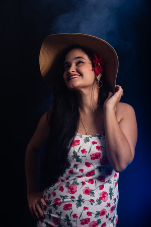 Beautiful smiling young woman with black hair, standing, wearing a brown beach hat with a red flower in her left ear wearing colorful clothes. Isolated on dark smoky background.の写真素材