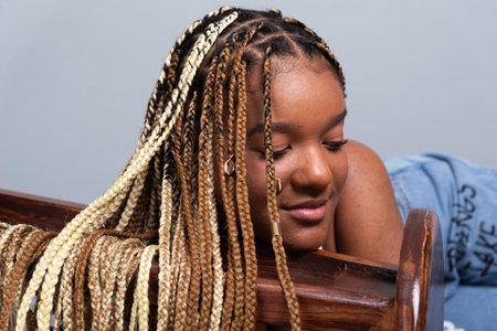 Portrait of a smiling brunette woman with braids in her hair, sitting on the floor in a studio leaning on a wooden stool. Isolated on a gray background.の写真素材
