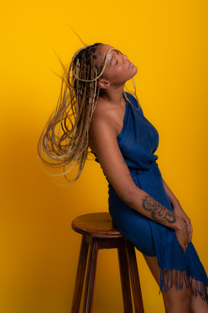 Photo of beautiful young brunette woman with braids in her hair wearing blue clothes, standing, leaning on a wooden stool posing for the camera. Isolated on yellow background.の写真素材