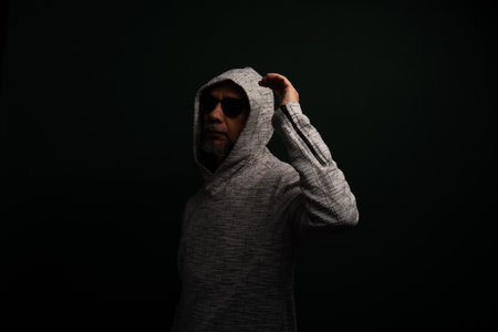 Portrait of man wearing sunglasses and hooded sweatshirt standing, motionless, making hand gestures. Isolated on dark background.の写真素材