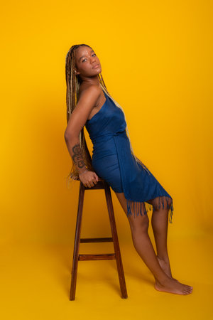 Photo of beautiful young brunette woman with braids in her hair wearing blue clothes, standing, leaning on a wooden stool posing for the camera. Isolated on yellow background.の写真素材