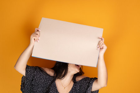 Beautiful black haired latin american woman holding blank banner above her head, smiling and confident, standing. Isolated on mustard colored background.の写真素材
