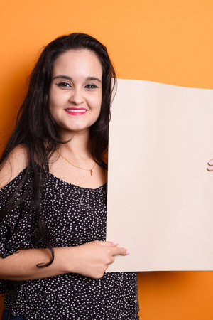 Young latin american woman holding a blank banner with a happy face, standing with a confident smile, pointing at the billboard and looking at the camera. Isolated on mustard colored background.の写真素材