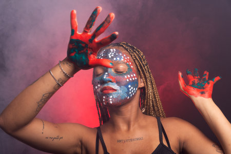 Woman with face and hands painted with colorful paint and braids in her hair, eyes closed, making gestures with hands and fingers. Isolated on background with red smoke.の写真素材