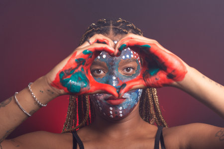 Woman with colorful face and hands paints and braids in her hair making a heart gesture in front of her eyes with her hands. Isolated on a background with red smoke.の写真素材
