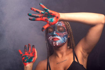 Woman with face and hands painted with colorful paint and braids in her hair, eyes closed, making gestures with hands and fingers. Isolated on background with red smoke.の写真素材