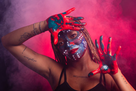 Woman with face and hands painted with colorful paint and braids in her hair making gestures with her hands and fingers. Isolated on a background with red smoke.の写真素材