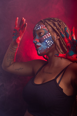 Woman with face and hands painted with colorful paint and braids in her hair making gestures with...