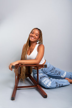 Beautiful brunette woman with braids in her hair sitting on the studio floor leaning on a wooden stool wearing denim clothes. Calm and confident. Isolated on gray background.の写真素材