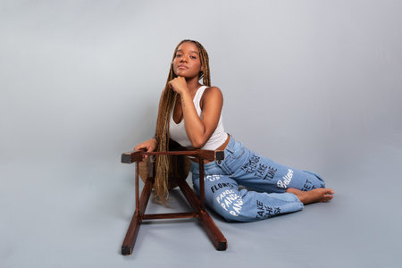 Beautiful brunette woman with braids in her hair sitting on the studio floor leaning on a wooden stool wearing denim clothes. Calm and confident. Isolated on gray background.の写真素材