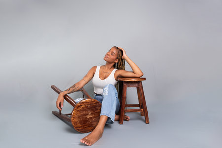 Beautiful brunette woman with braids in her hair, sitting on the floor of a studio leaning on a wooden stool. Calm and confident thoughtful person. Isolated on a gray background.の写真素材