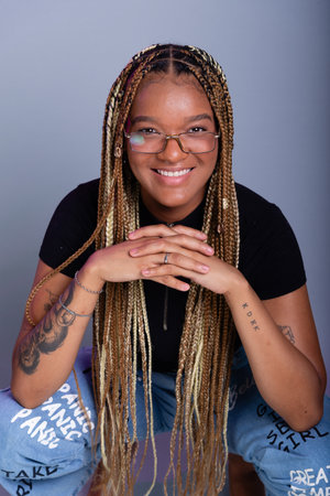 Brunette model with braids in her hair, wearing prescription glasses, a denim dress and a black blouse, crouching on the studio floor. Isolated on a gray background.の写真素材