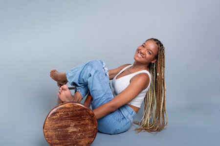 Portrait of a smiling brunette woman with braids in her hair, sitting on the floor in a studio leaning on a wooden stool. Isolated on a gray background.の写真素材