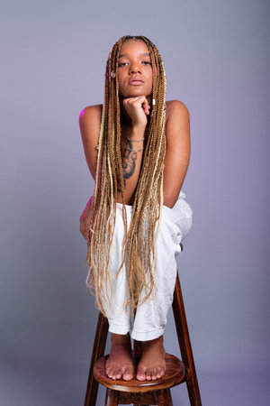 Brunette model with braids in her hair, wearing white jeans and a black blouse sitting on a wooden stool posing for the camera. Isolated on a gray background.の写真素材