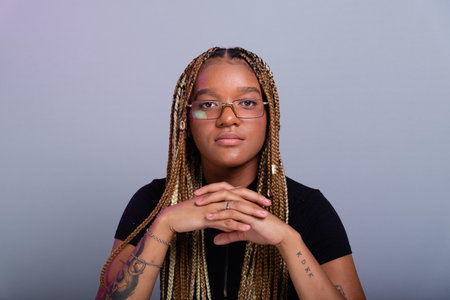 Brunette model with braids in her hair, wearing prescription glasses, a black blouse, crouching on the studio floor. Isolated on a gray background.の写真素材