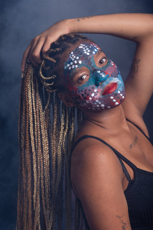 Beautiful young woman with colorful face paint holding her hair braids and posing for the camera. Isolated on dark smoky background.の写真素材