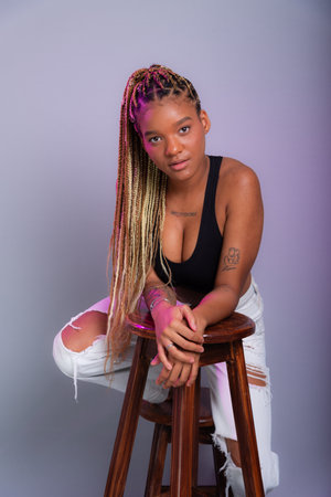 Model with braids in her hair, wearing white jeans and a black top leaning on a wooden stool posing for the camera. Isolated on a gray background.の写真素材