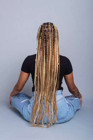 Brunette woman with braids in her hair, wearing jeans and a black blouse, sitting on the studio floor with her back to the camera. Isolated on a gray background.の写真素材