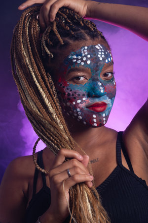 Beautiful young woman with colorful face paint holding her hair braids and posing for the camera. Isolated on dark smoky background.の写真素材
