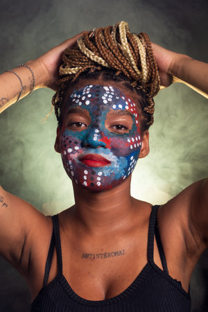 Woman with face painted with colorful paint and braids in her hair with her hands on her head. Isolated on a background with light green smoke.の写真素材