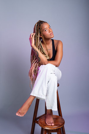 Brunette model with braids in her hair, wearing white jeans and a black blouse sitting on a wooden stool posing for the camera. Isolated on a gray background.の写真素材