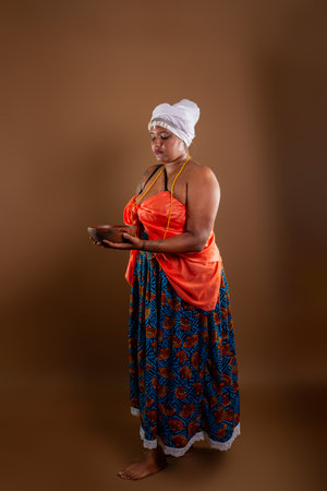 Salvador, Bahia, Brazil - October 21, 2024: Beautiful black woman wearing traditional Candomble clothing, holding a clay pot against brown background. Salvador, Bahia.の写真素材