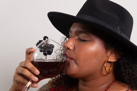 Salvador, Bahia, Brazil - October 21, 2024: Beautiful woman in black hat drinking wine from glass cup with black grapes. isolated on white background.の写真素材
