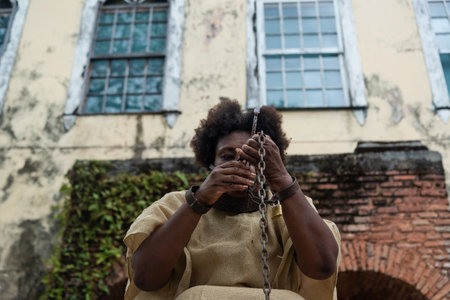 Black woman dressed as a slave, chained and with an iron mask on her mouth and neck, crouched against a colonial house. Slavery in Brazil. Expression of pain and sadnessの写真素材