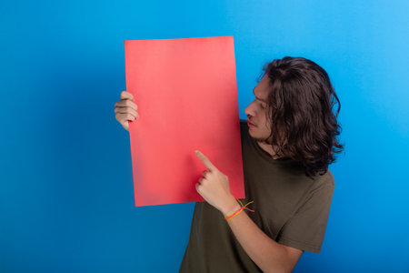 young man holding an empty red colored poster. Confident and pointing finger to the center of the poster. Isolated on blue background.の写真素材