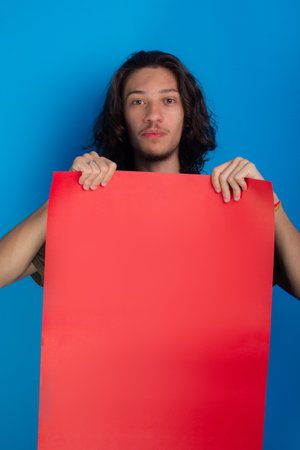 young man holding an empty red colored poster. Confident and looking at camera. Isolated on blue background.の写真素材