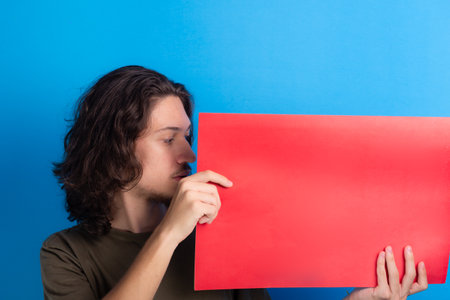 young man holding an empty red colored poster. Confident and looking at the poster. Isolated on blue background.の写真素材