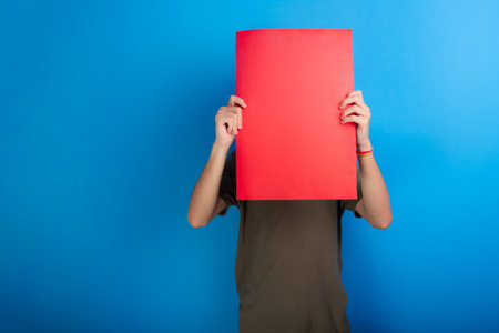 young man holding empty red colored poster in front of face. Isolated on blue background.の写真素材