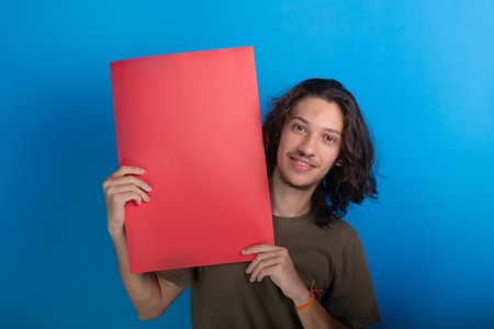 young man holding an empty red colored poster. Confident and looking at camera. Isolated on blue background.の写真素材