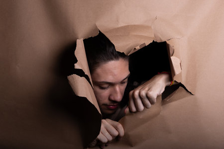 Man's head sticking out and looking through a paper hole with his hands pushing the paper aside. Studio portrait.の写真素材