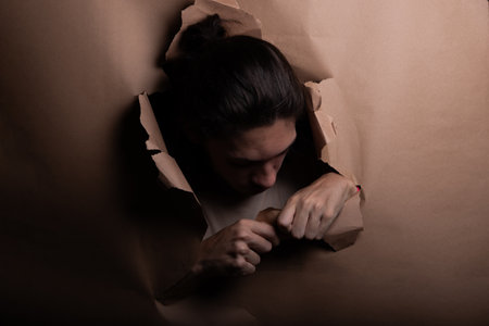 Man's head sticking out and looking through a paper hole with his hands pushing the paper aside. Studio portrait.の写真素材