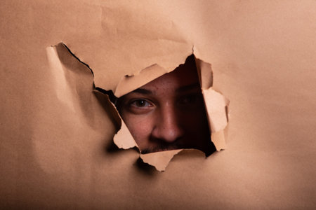 Man looking through a paper hole with his hands pushing the paper aside. Studio portrait.の写真素材