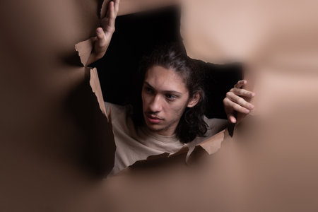 Man's head sticking out and looking through a paper hole with his hands pushing the paper aside. Studio portrait.の写真素材