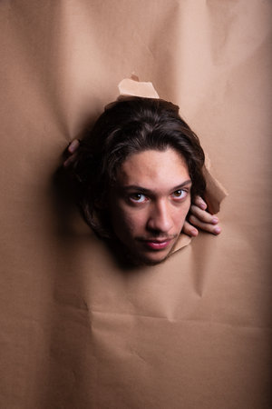 Man's head sticking out and looking through a paper hole with his hands pushing the paper aside. Studio portrait.の写真素材