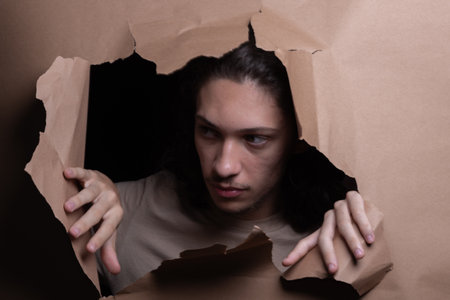 Man's head sticking out and looking through a paper hole with his hands pushing the paper aside. Studio portrait.の写真素材