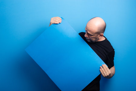 Bearded, bald man in black shirt holding and looking at blue poster. Studio shot.の写真素材