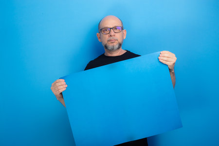 Bearded, bald man wearing prescription glasses and a black shirt holding a blue poster. Confident and focused. Studio shot.の写真素材