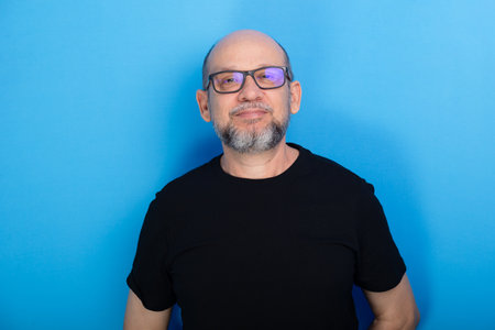 Bearded, bald man wearing prescription glasses and a black shirt posing for a photo against a blue...