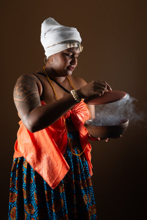 Salvador, Bahia, Brazil - October 21, 2024: Beautiful woman in traditional Candomble clothing, opening clay pot with smoke coming out of it. Salvador, Bahia.の写真素材