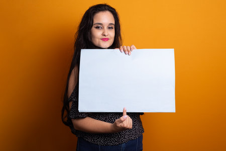 Portrait of beautiful black haired woman holding blank poster and looking at camera. Isolated on yellow background.の写真素材