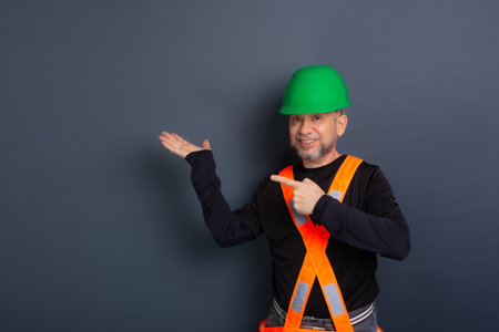 A civilian worker wearing a green helmet, black clothing, reflective vest and pointing with his finger to the side. isolated on gray background. Construction worker.の写真素材